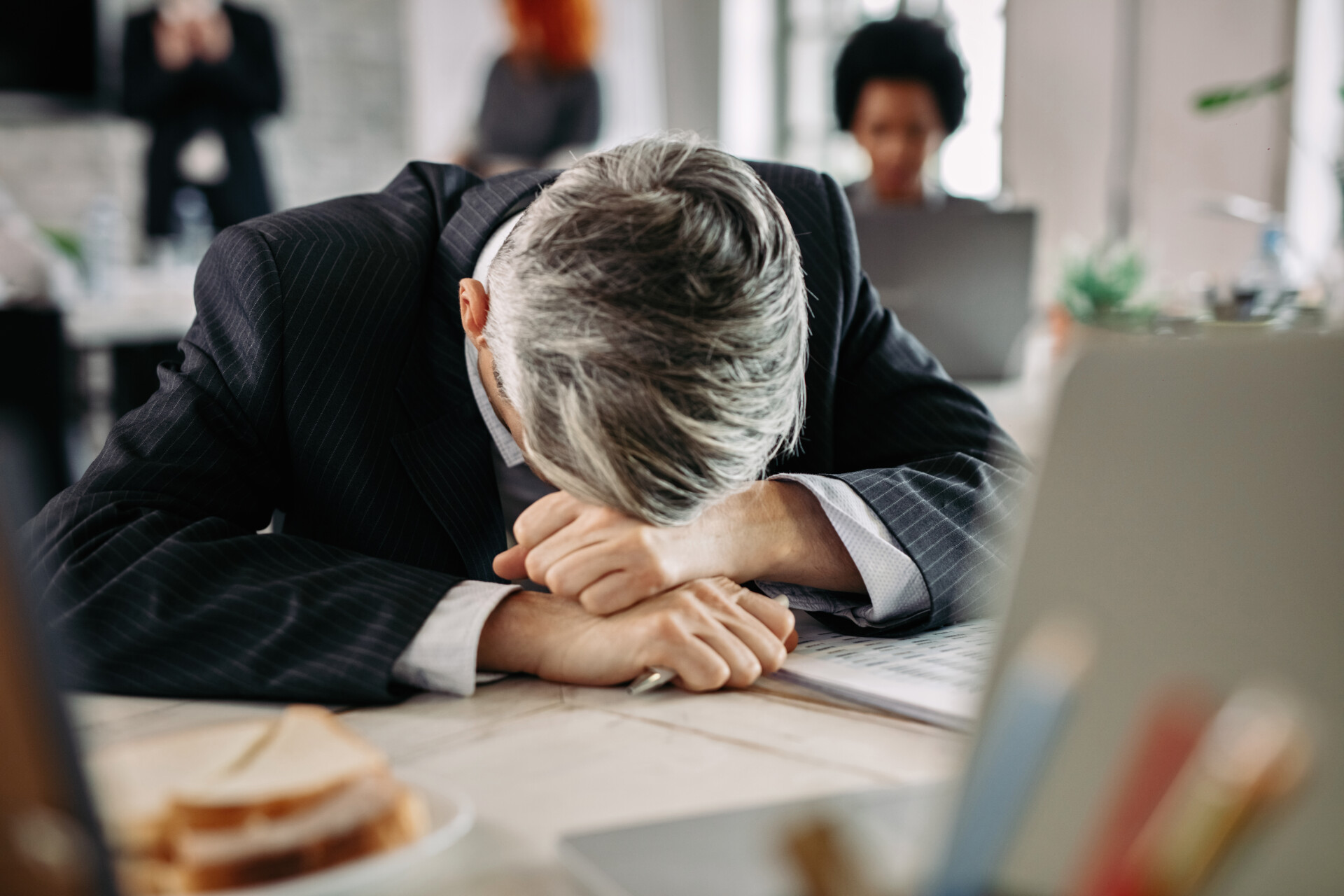 Overworked businessman feeling tired in the office and resting his head at the desk. There are people in the background.